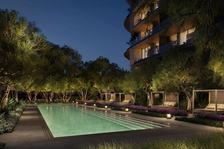 Rendu de la piscine des Aman Residences Beverly Hills la nuit, bordée d'oliviers et de cabanas, au pied d'une tour du complexe One Beverly Hills