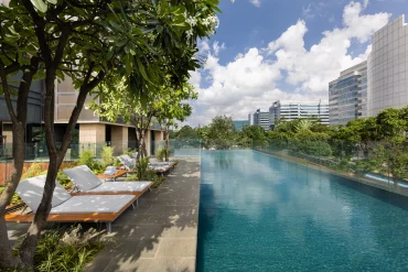 La piscine extérieure à débordement de The Den Bengaluru, LXR Hotels & Resorts, bordée de transats et de frangipaniers face aux tours de Whitefield