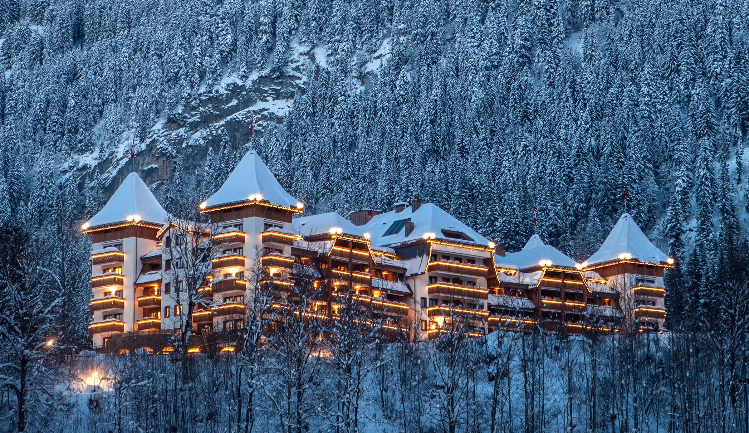 L'hôtel The Alpina Gstaad illuminé un soir d'hiver, ses tours coiffées de neige au-dessus de la forêt, à Gstaad en Suisse